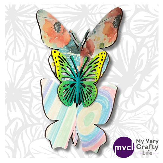 Photo Depicts a gray and white background of flower drawings. A set of 3, 3D Custom Made Butterflies, from the 3D Butterfly SVG Files,in various prints are shown stacked on one another. The logo in purple for "My Very Crafty Life" is in the bottom right corner